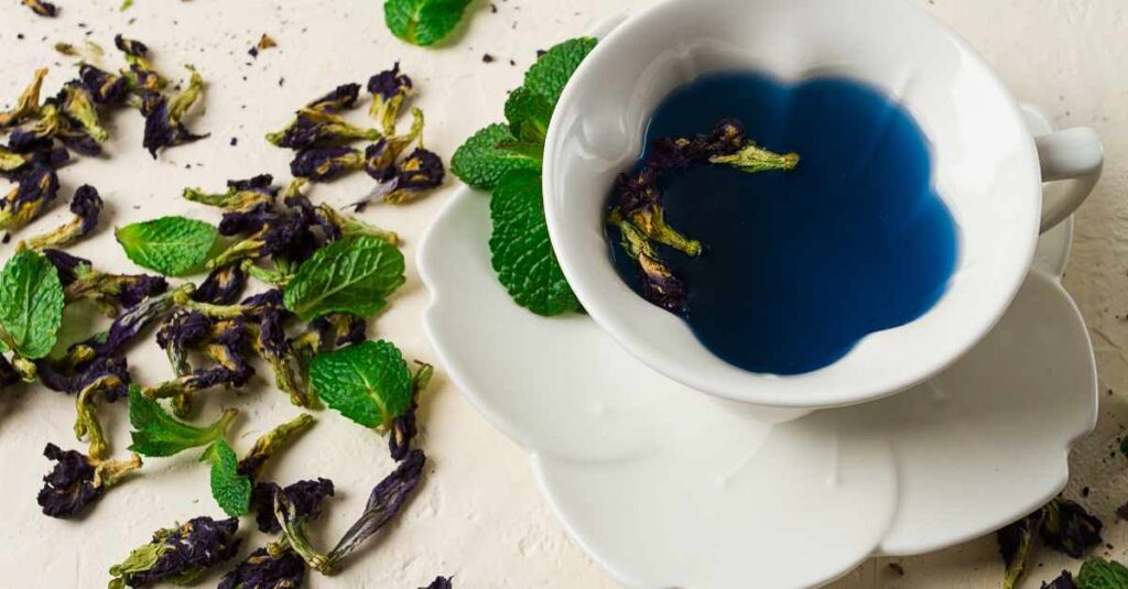 A white teacup filled with vibrant blue butterfly pea flower tea, garnished with dried butterfly pea flowers and fresh mint leaves, surrounded by scattered dried flowers and mint on a light surface